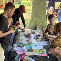 Kinder und Erwachsene sitzen gemeinsam an einem langen Tisch im Freien und basteln mit buntem Papier. Sie falten Papierboote, schneiden und malen; auf dem Tisch liegen Scheren, Stifte, Anleitungen und farbige Bastelmaterialien. Im Hintergrund sind Bäume, Fahrräder und Wimpel zu sehen.