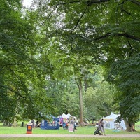 Weitläufige Parkwiese mit großen Bäumen und mehreren Zelten einer Veranstaltung im Hintergrund.