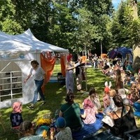 Eine große Gruppe von Kindern sitzt auf dem Gras und hört einer Lesung zu. Im Vordergrund ist eine Frau auf der Bühne zu sehen, die eine Geschichte vorträgt. Das Zelt im Hintergrund ist mit bunten Vorhängen geschmückt, und die Atmosphäre ist lebendig und festlich.